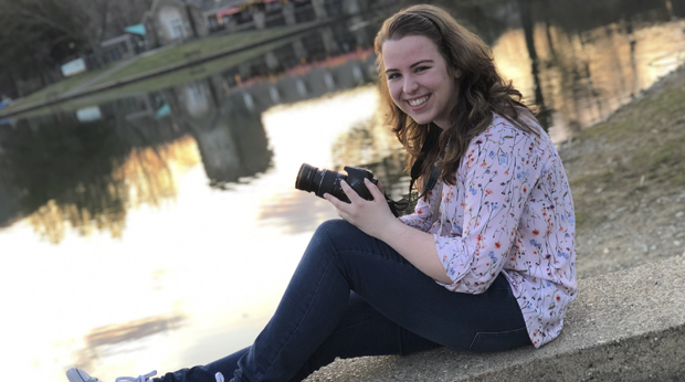 La Roche University student Sarah Hefferin sits in front of North Park Lake and Boathouse holding a camera.